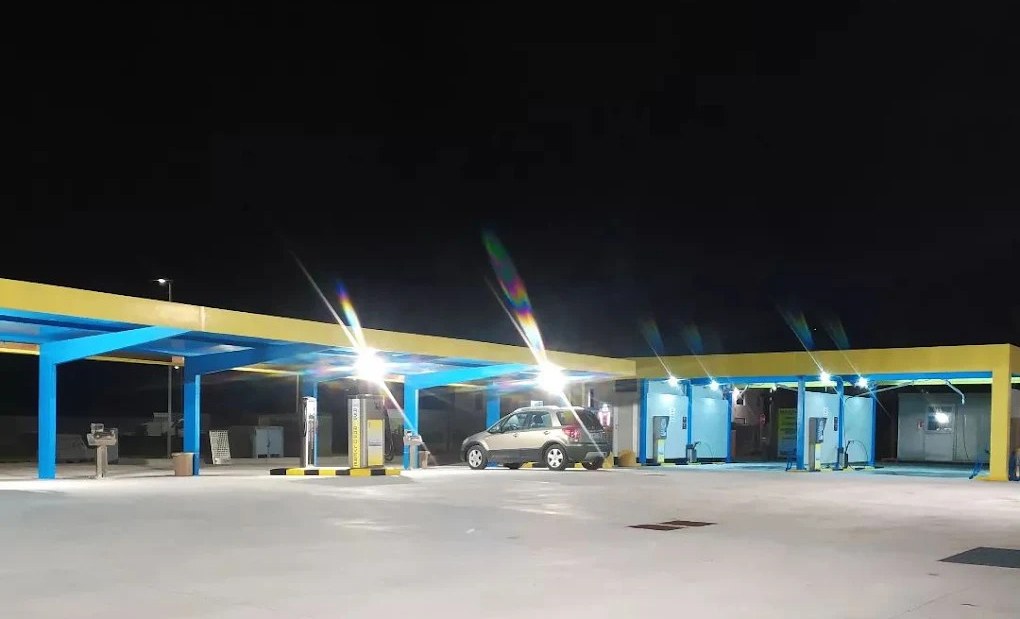 Autolavaggio self service di notte con pensiline gialle e blu illuminate, auto grigia in sosta e piazzale vuoto